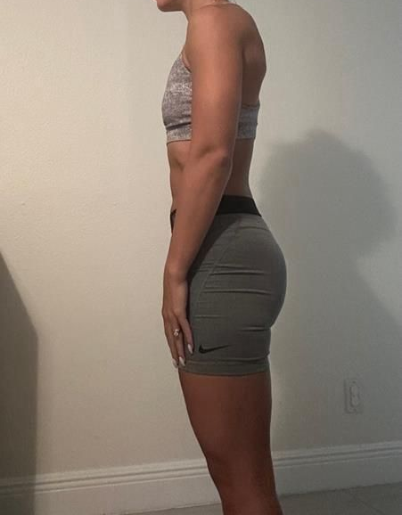 Woman in athletic wear, standing sideways, showing physique against a plain wall.