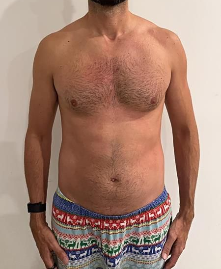 Man with a hairy chest wearing festive pajama pants, standing with hands in pockets.
