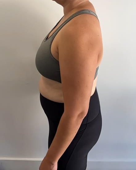 Woman in a sports bra and leggings, standing in profile against a plain wall.