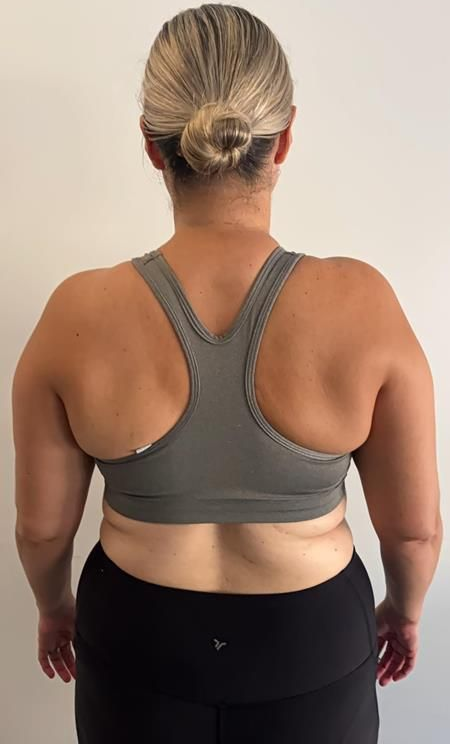 Woman from the back, wearing a gray sports bra and black leggings, standing against a white wall.
