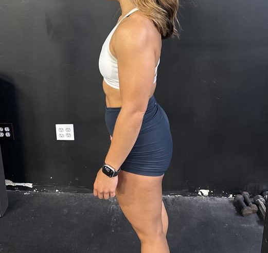 Woman in workout clothes, standing sideways in gym, looking down. Black wall background.