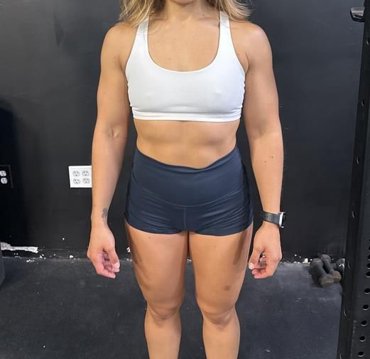 Woman in white sports bra and shorts stands in front of a black wall, a watch on her left wrist.