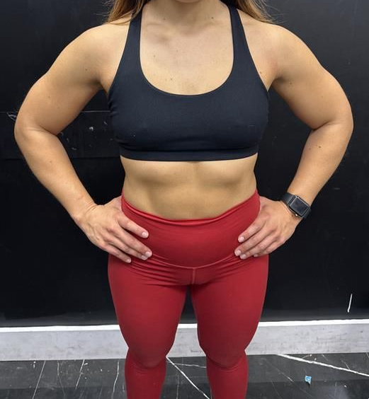 Woman in workout attire, hands on hips, arms flexed, in front of a black wall.