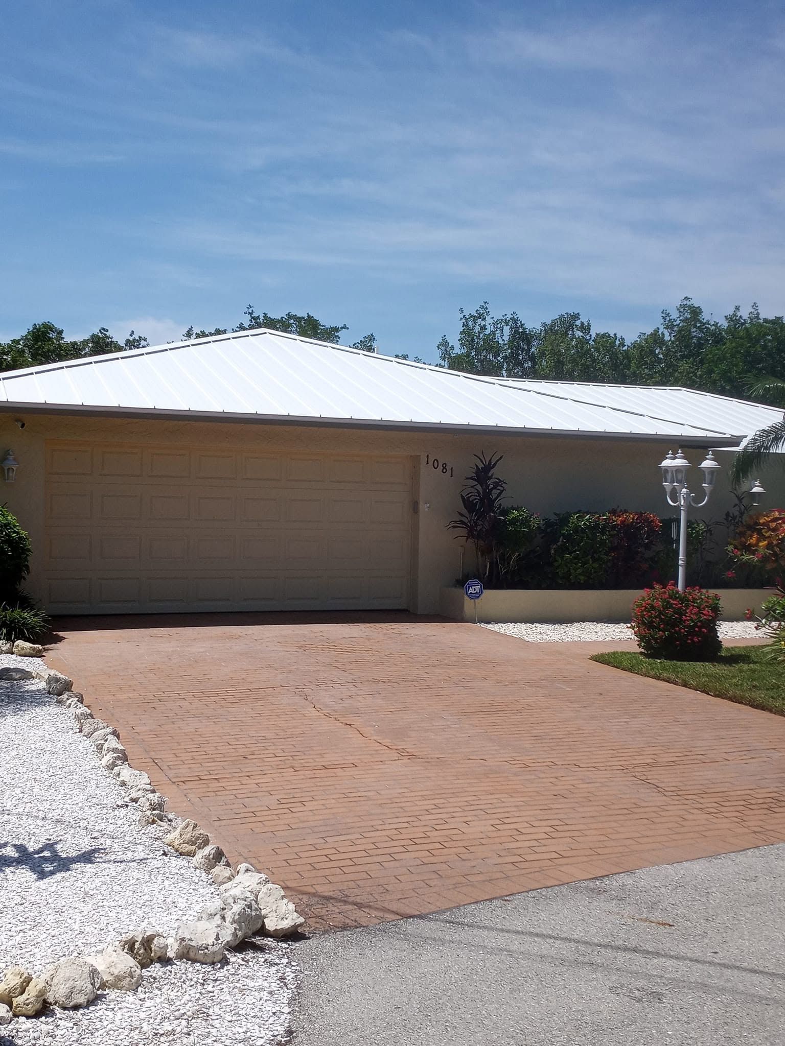Single-story house with a white roof, tan garage door, and brick driveway under a clear blue sky
