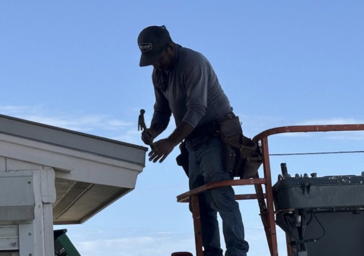 Worker on a lift repairing a roof edge beside a house, wearing safety gear.