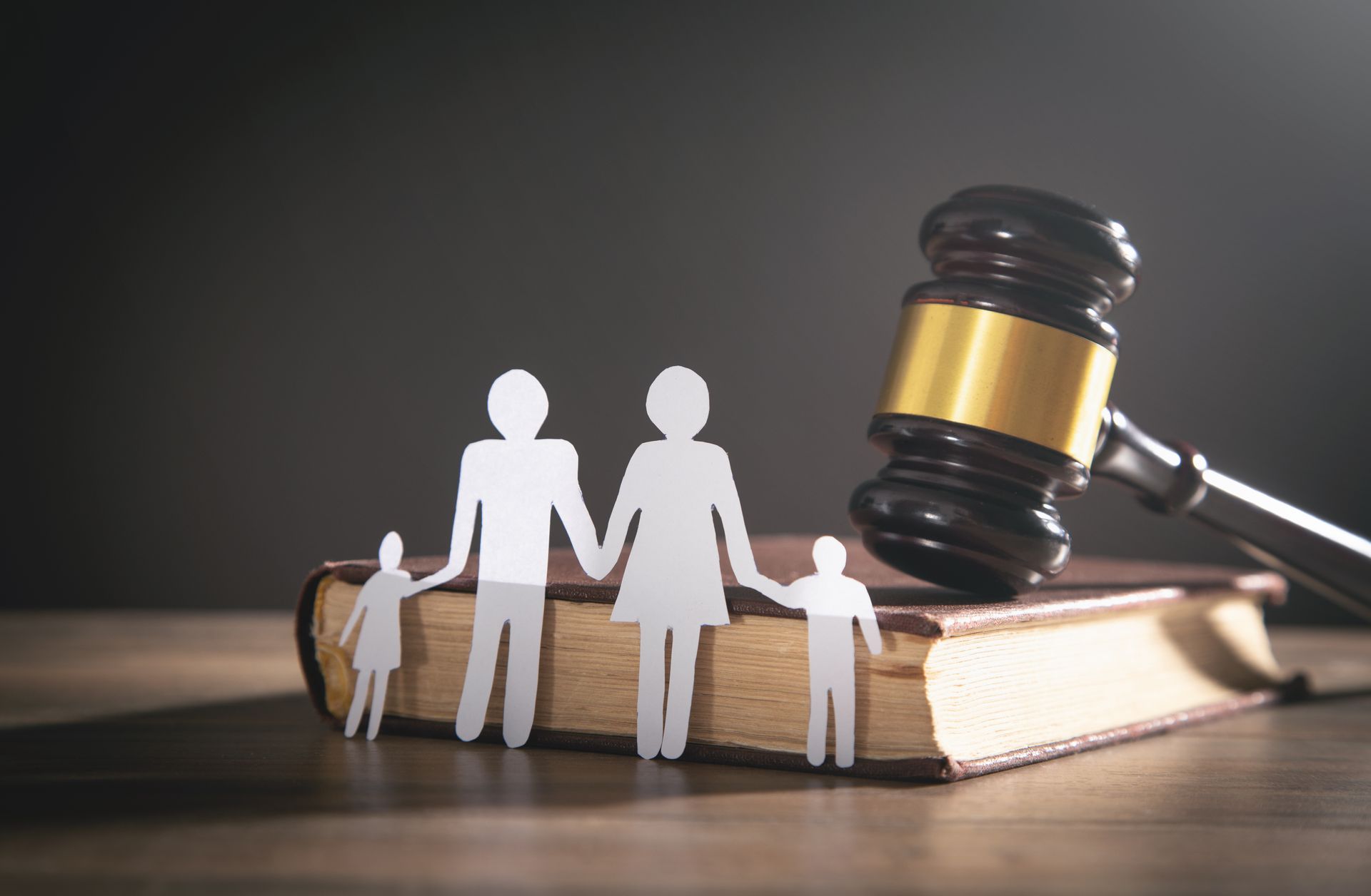 Paper cutout family figures standing on a closed law book with a judge’s gavel in the background.