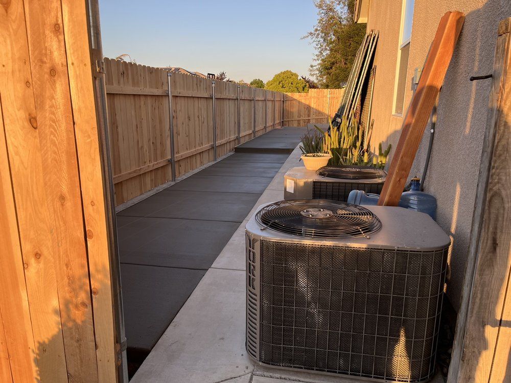 Two air conditioners are sitting on a sidewalk next to a wooden fence.