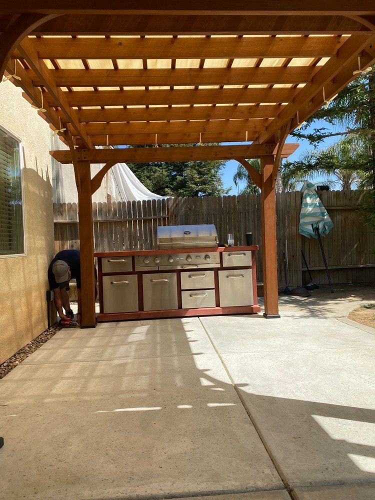 A man is working on an outdoor kitchen under a wooden pergola.