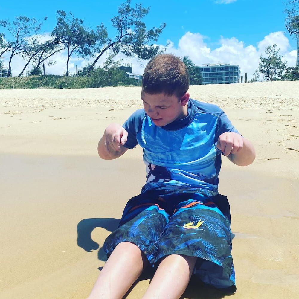 A Young Boy Wearing A Blue Shirt And Blue Shorts Is Sitting On The Beach — Tailored Support Services Pty Ltd In Bli Bli, QLD