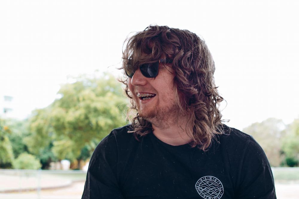 A Man With Long Curly Hair And Sunglasses Is Smiling — Tailored Support Services Pty Ltd In Maroochydore, QLD