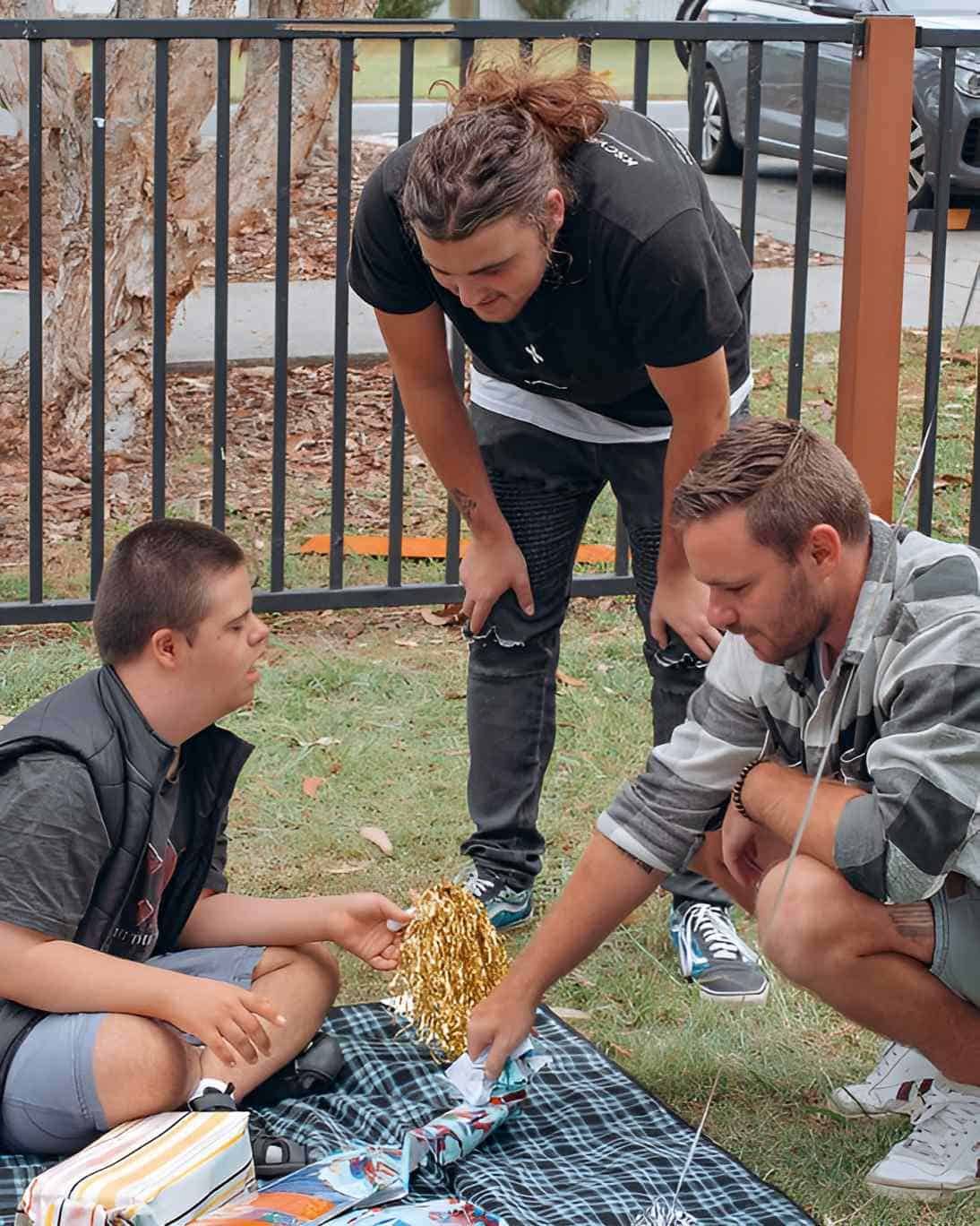 Three Men Are Sitting On A Blanket In The Grass Talking To Each Other — Tailored Support Services Pty Ltd In Maroochydore, QLD