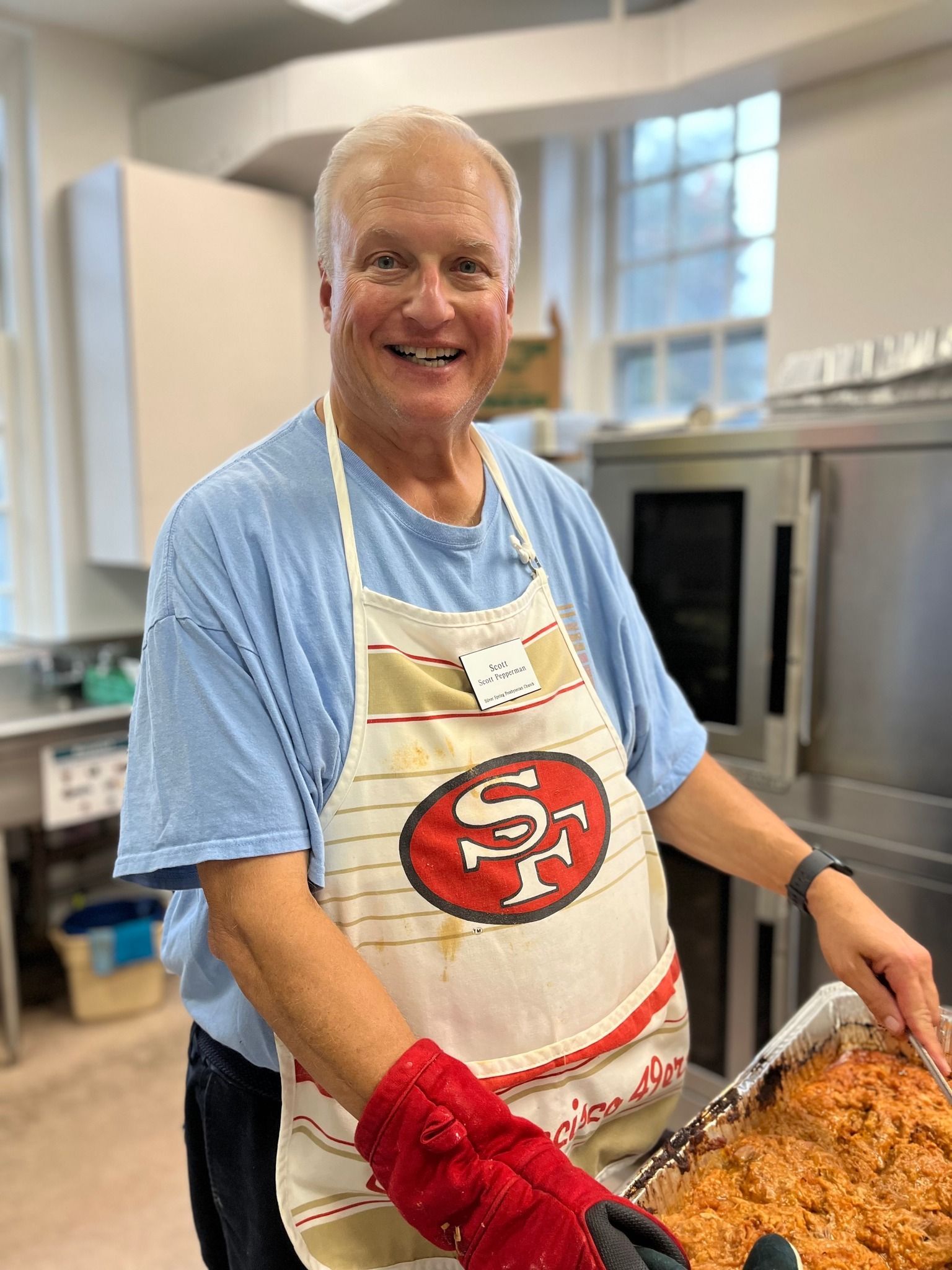 Smiling man wearing a 49ers apron, holding food, in a kitchen.