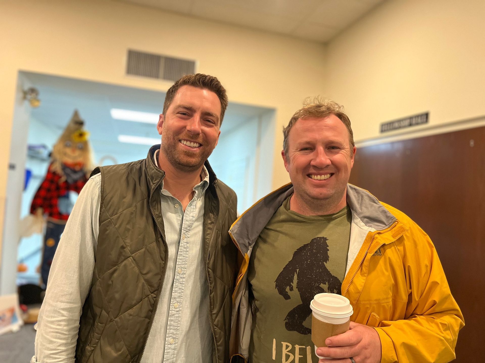 Two men smiling, one in a vest, the other holding a coffee with a Bigfoot shirt, in a hallway with a scarecrow.