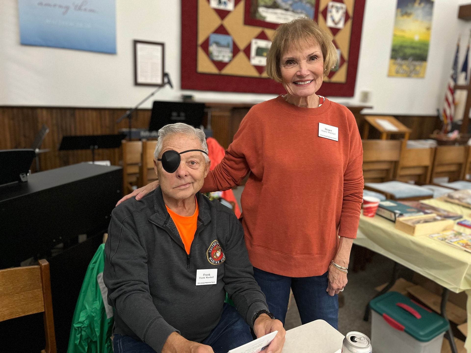 Two people smiling, one wearing an eye patch. A woman with her hand on the man's shoulder. Inside a church.