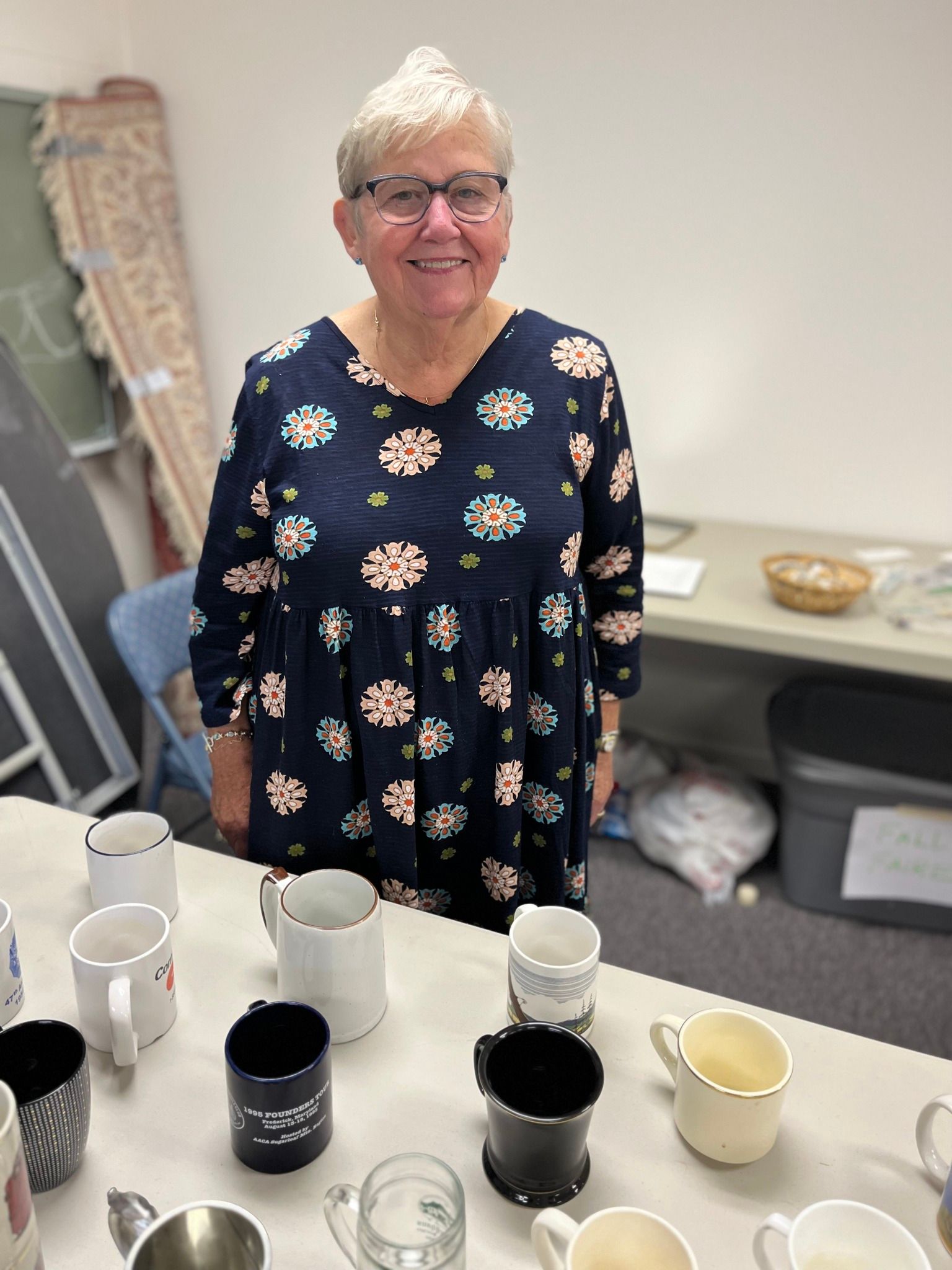 Woman smiles behind a table displaying various mugs. She wears glasses and a patterned dress.