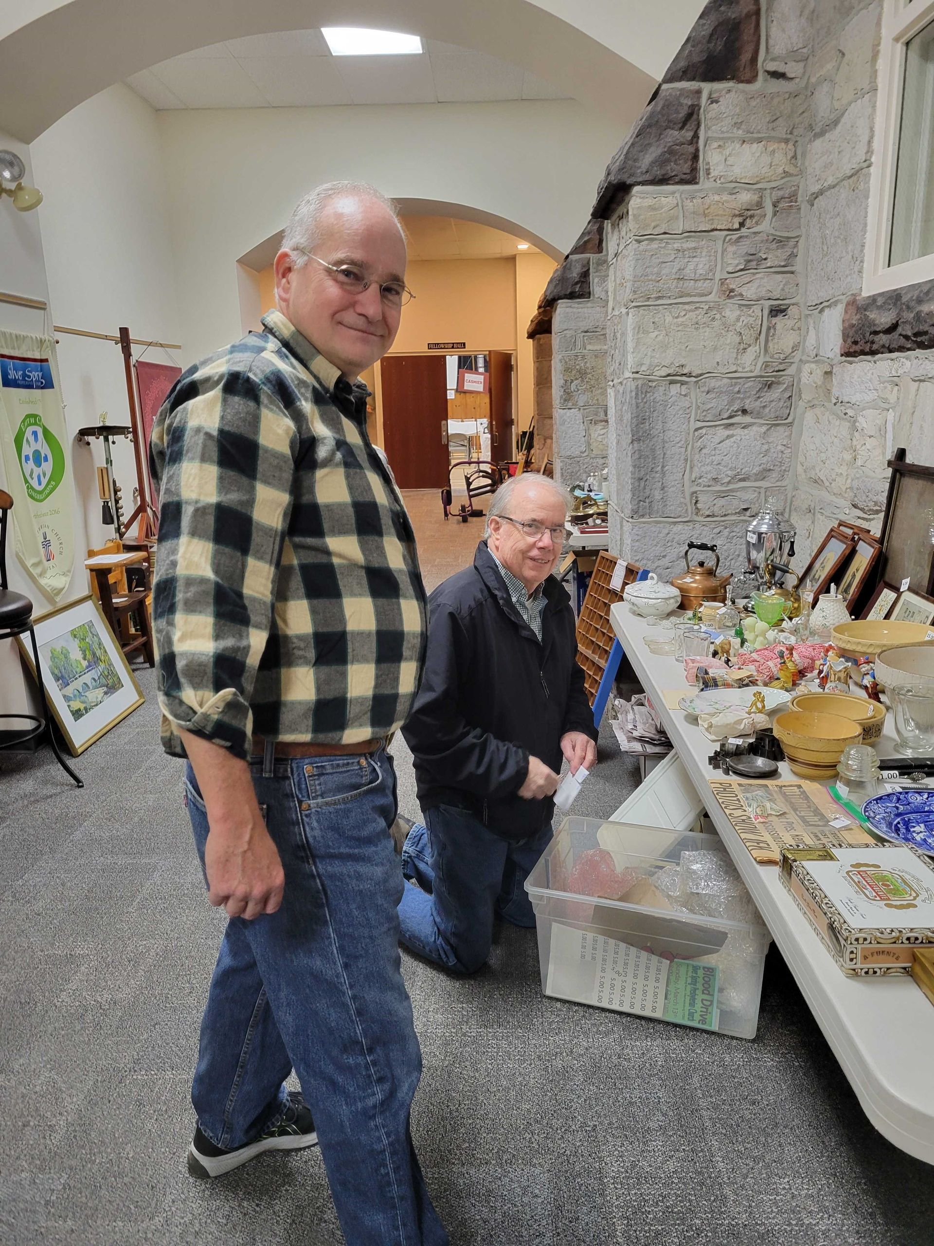 Two men browsing items at a table in a brightly lit room. One man is kneeling, another is smiling.