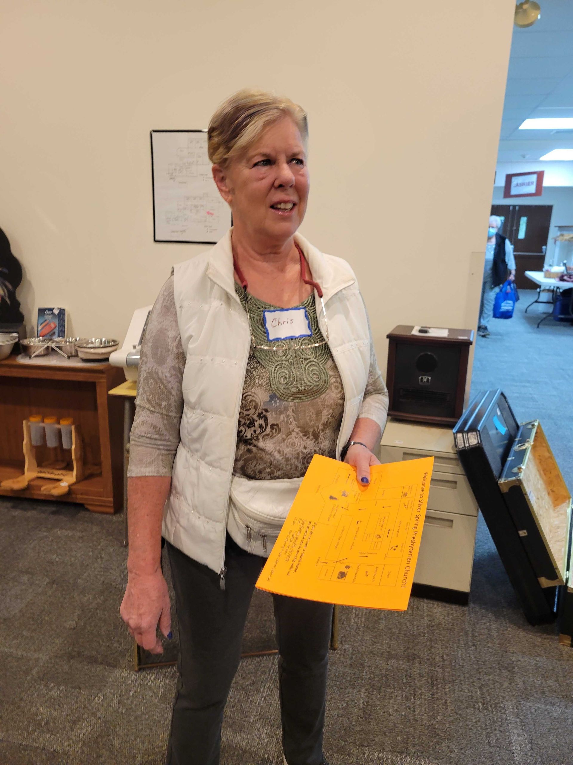 Woman in white vest and patterned top holds orange paper, indoors.