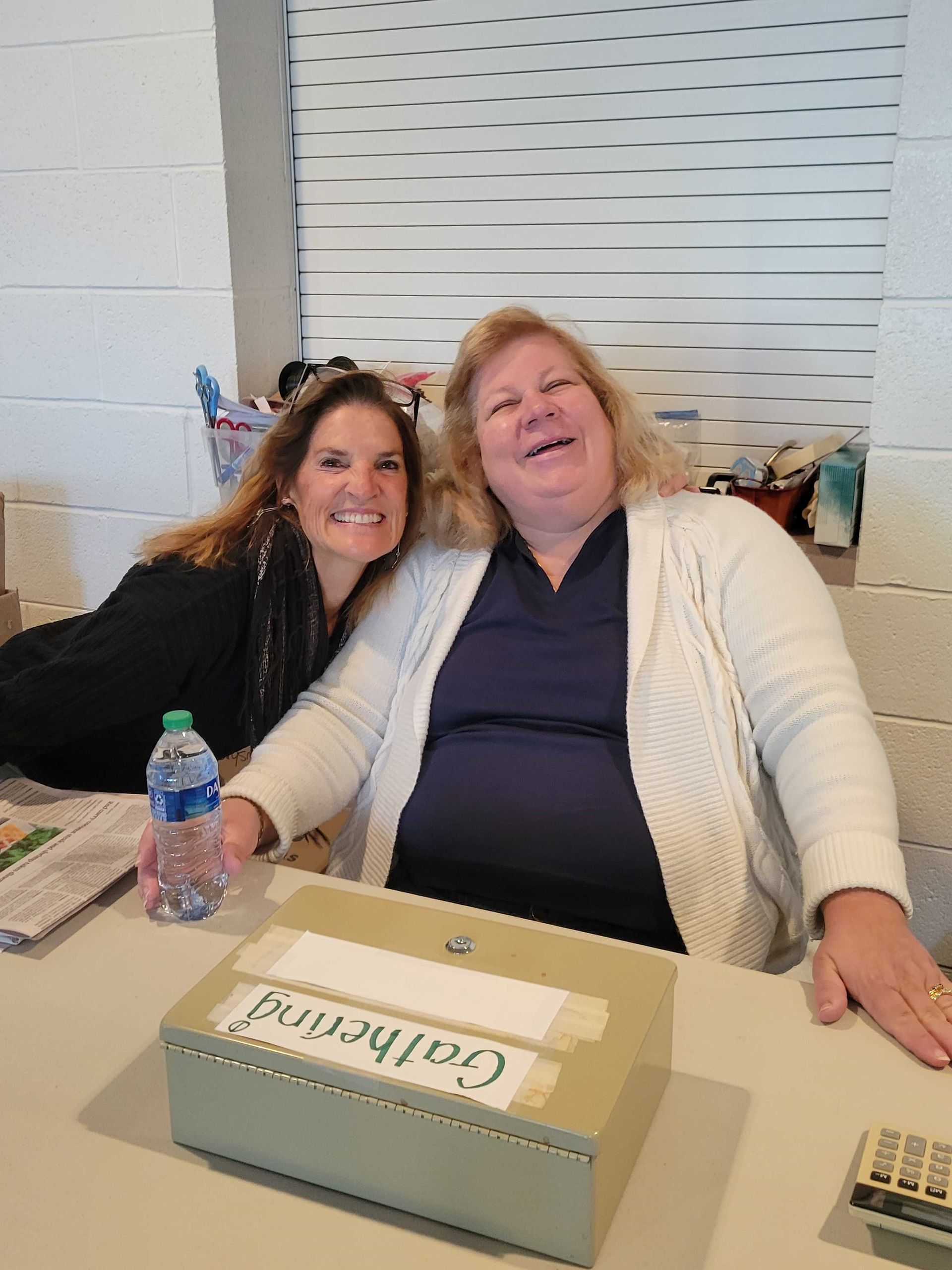 Two women smiling at a table. One wears a white sweater. A box labeled