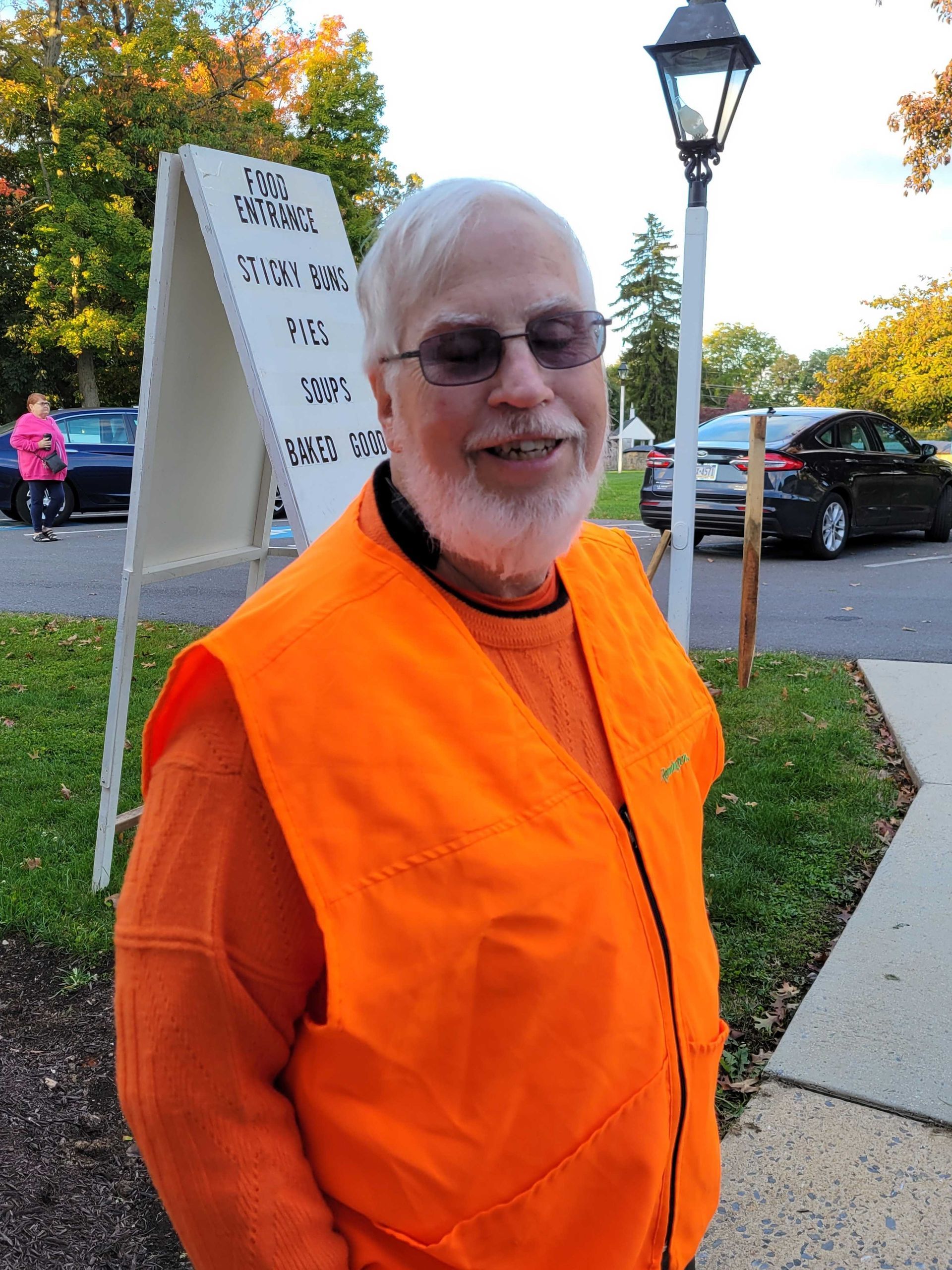 Man wearing orange vest and glasses smiles outside. Sign and cars in background.