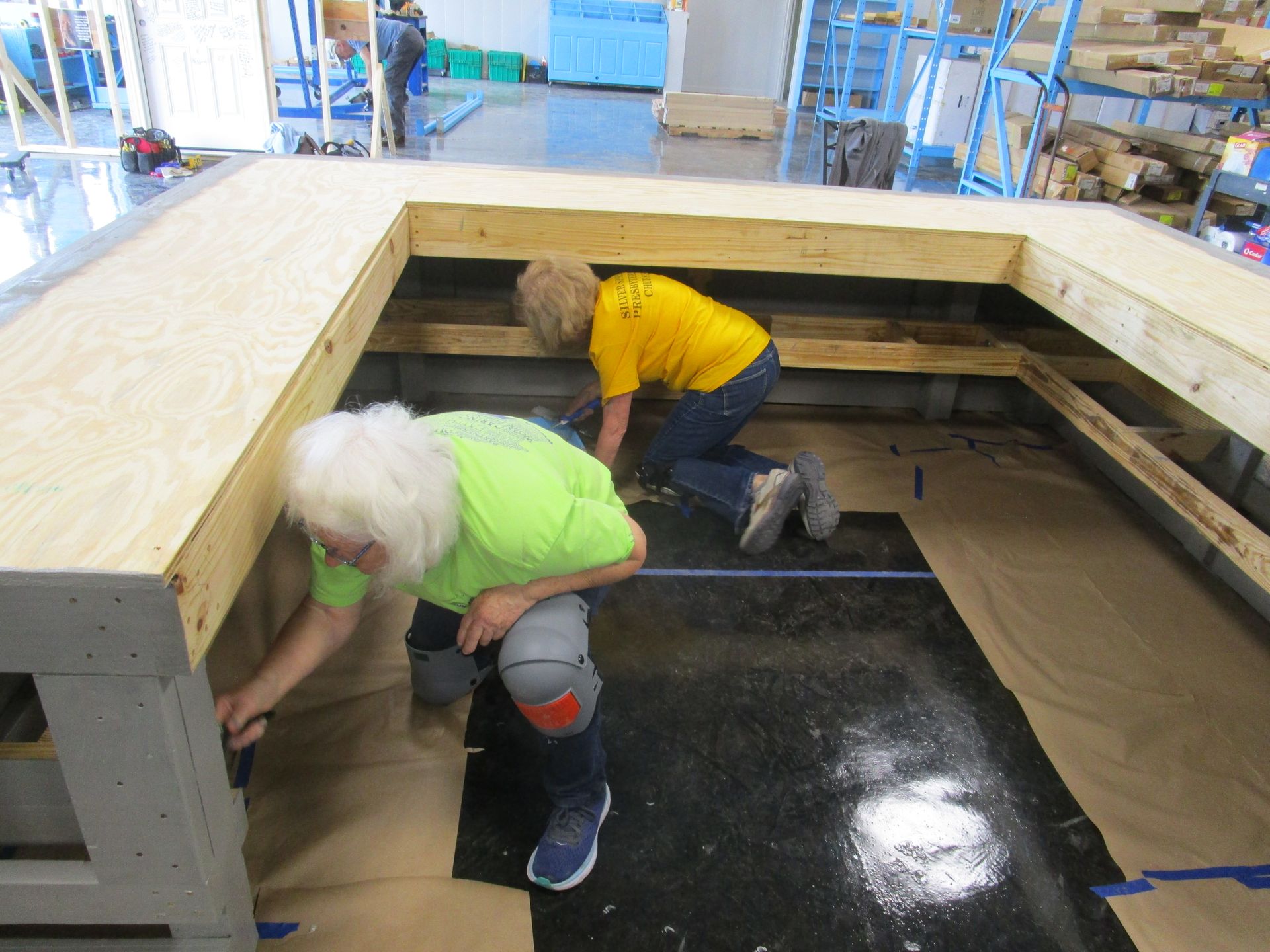 Two people installing a wooden structure indoors, with one kneeling and one bent over, both wearing work clothes.