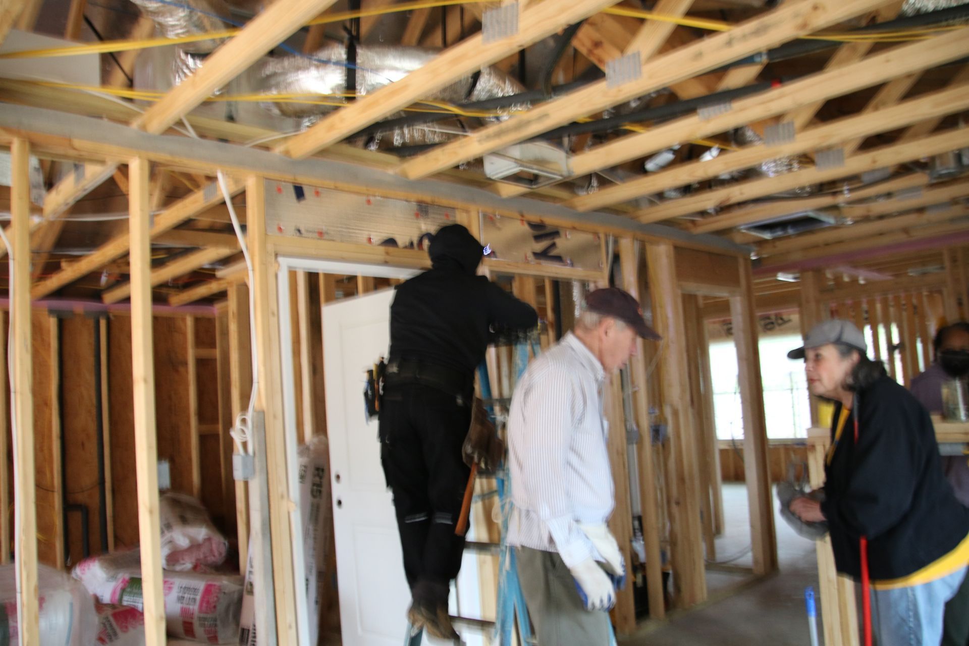 Construction workers inside a building frame. Man on ladder, others observing, working on framing, wiring visible.
