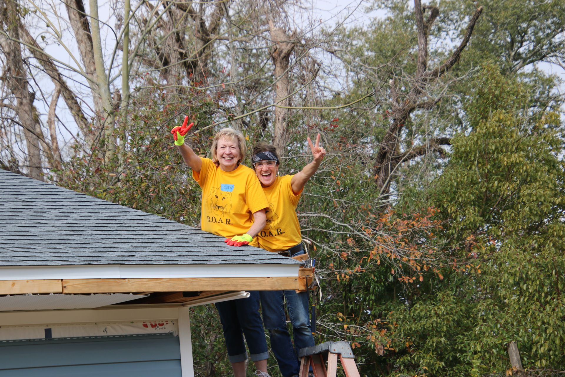 Two women on a roof, wearing yellow shirts and waving, with trees in the background.