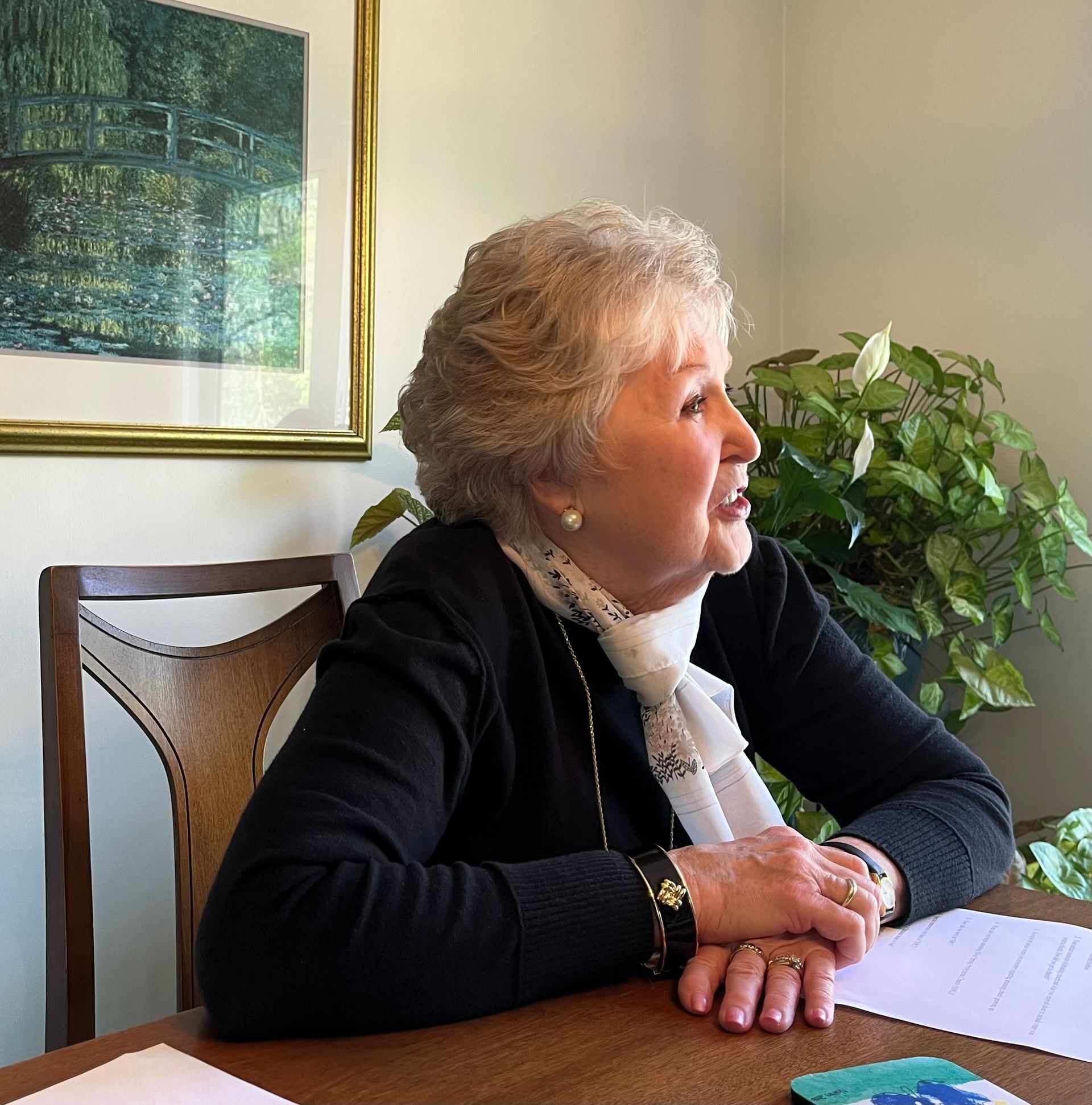 A woman sits at a table with a picture on the wall behind her
