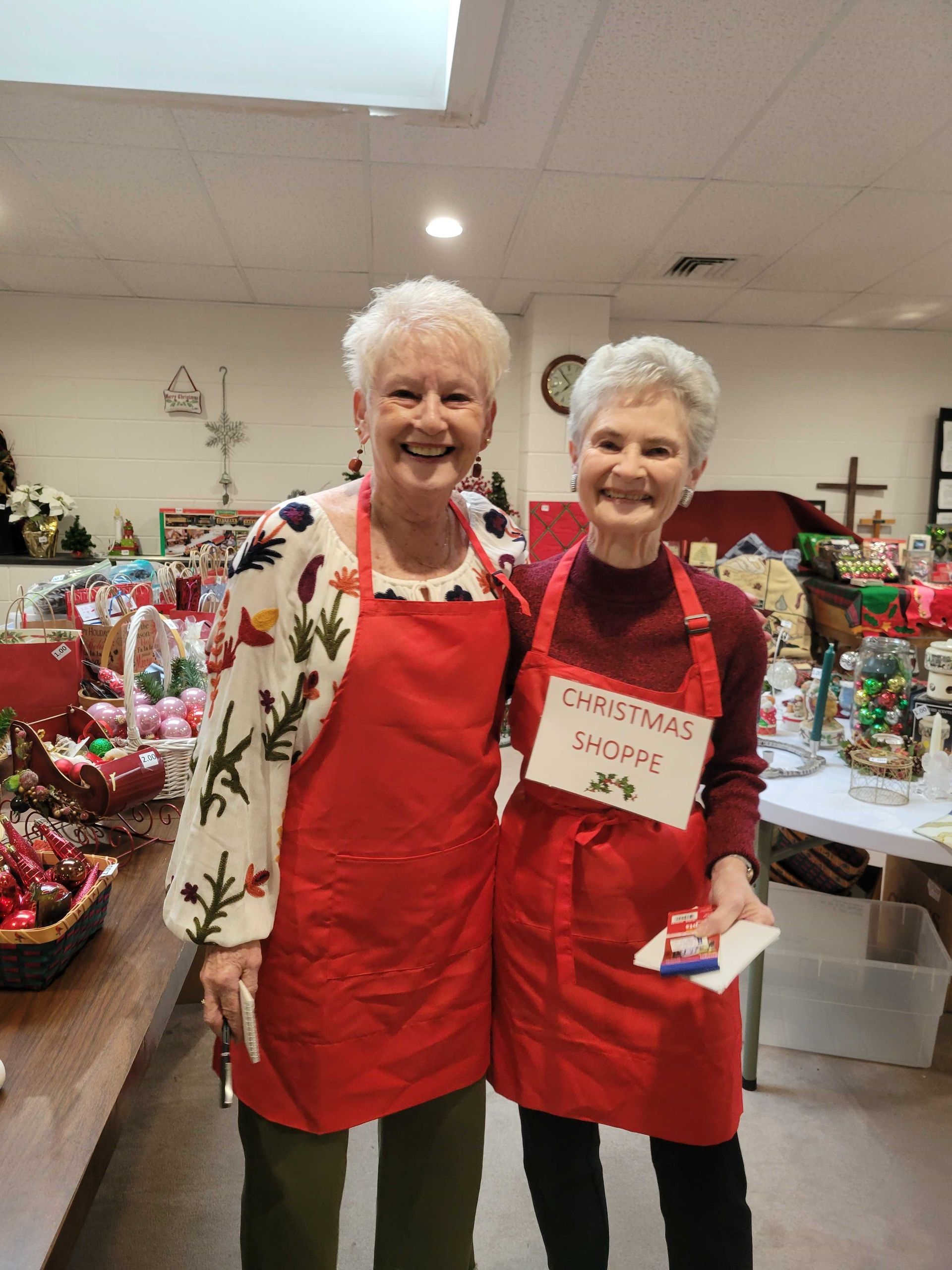 Two smiling women in red aprons at a Christmas shop.