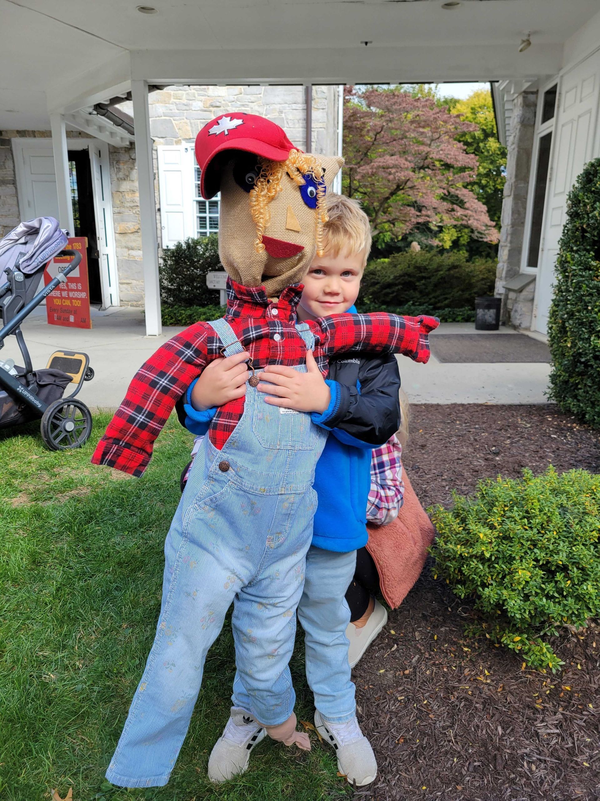 Young child hugging a scarecrow costume outdoors; both are smiling.