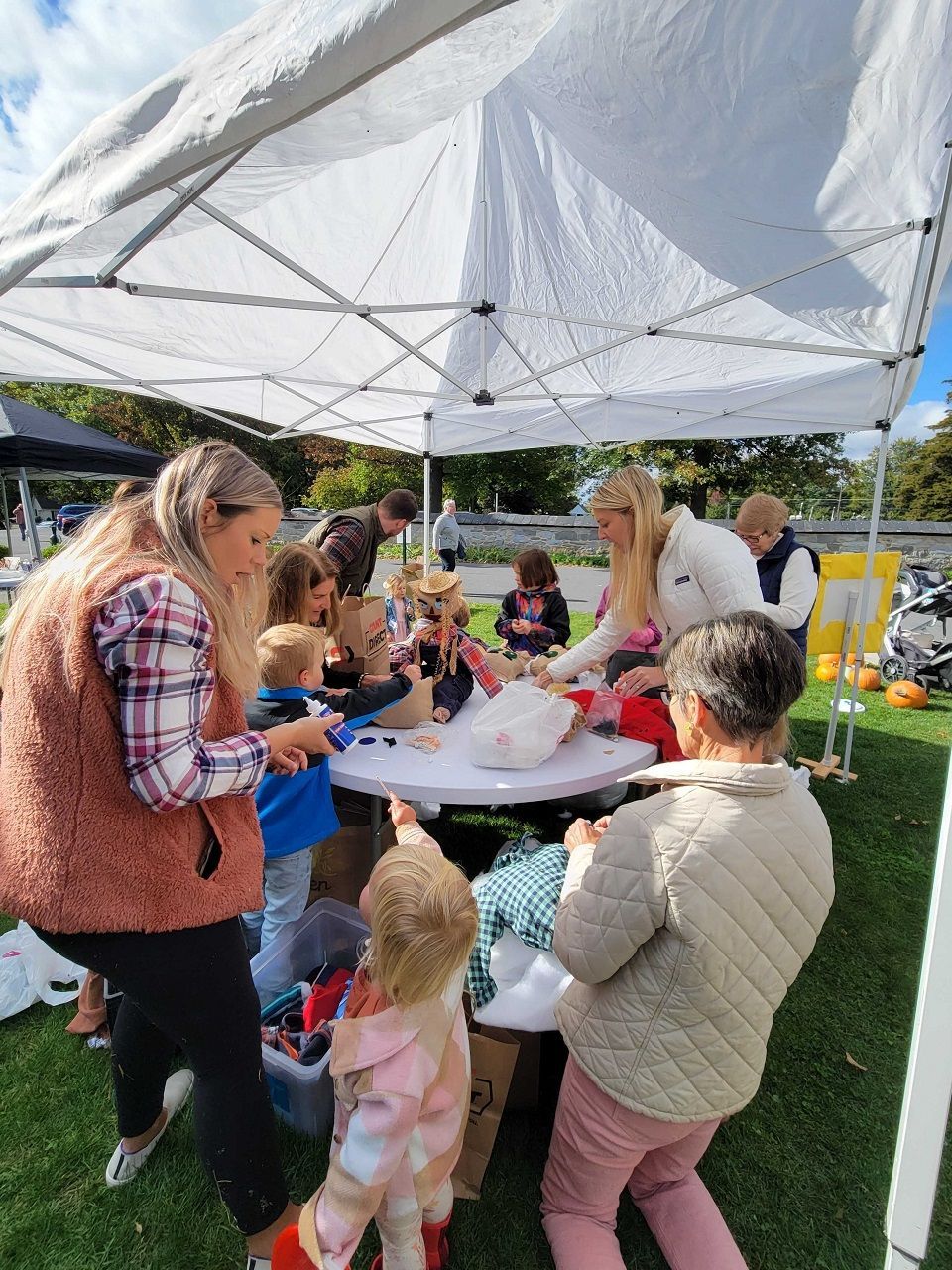 Children and adults at a craft table under a canopy, creating art outdoors.