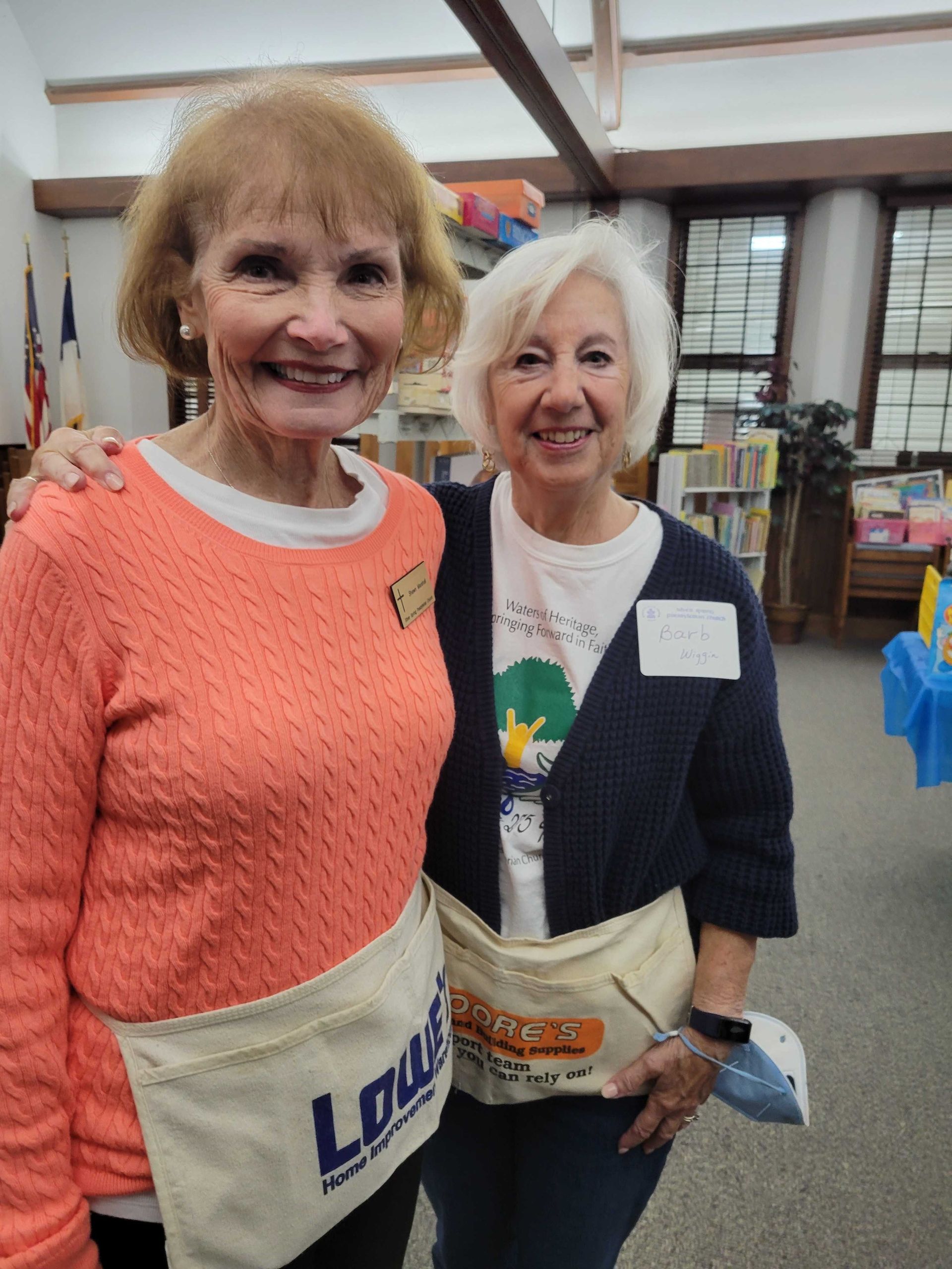 Two women smiling, wearing Lowe's aprons, in a library. One has her arm around the other.