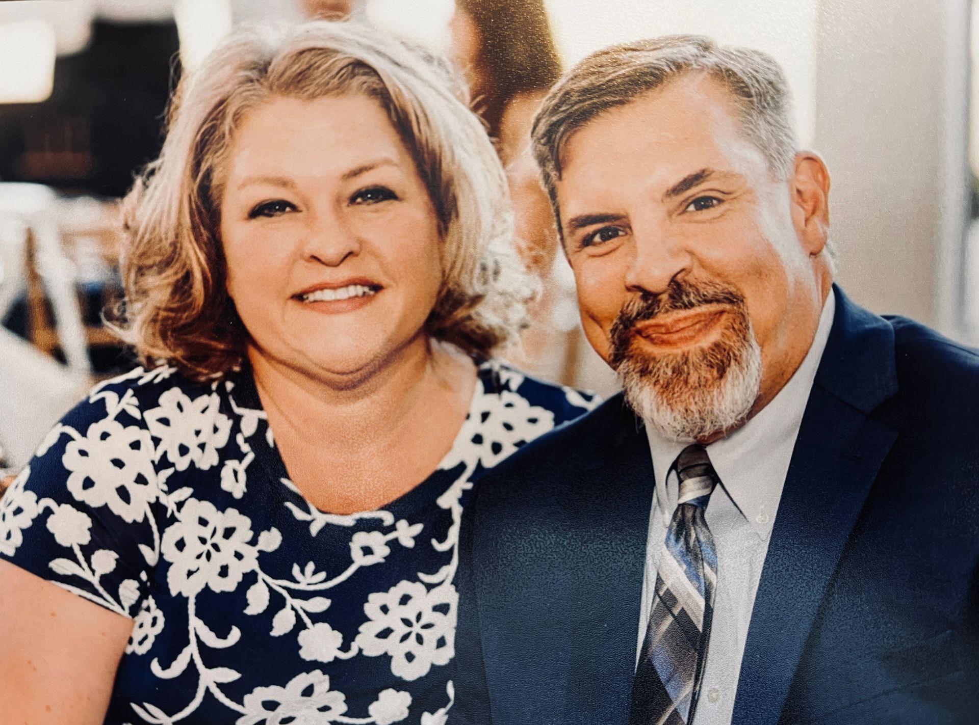 Couple smiling, woman in floral top, man in suit, outdoor setting.