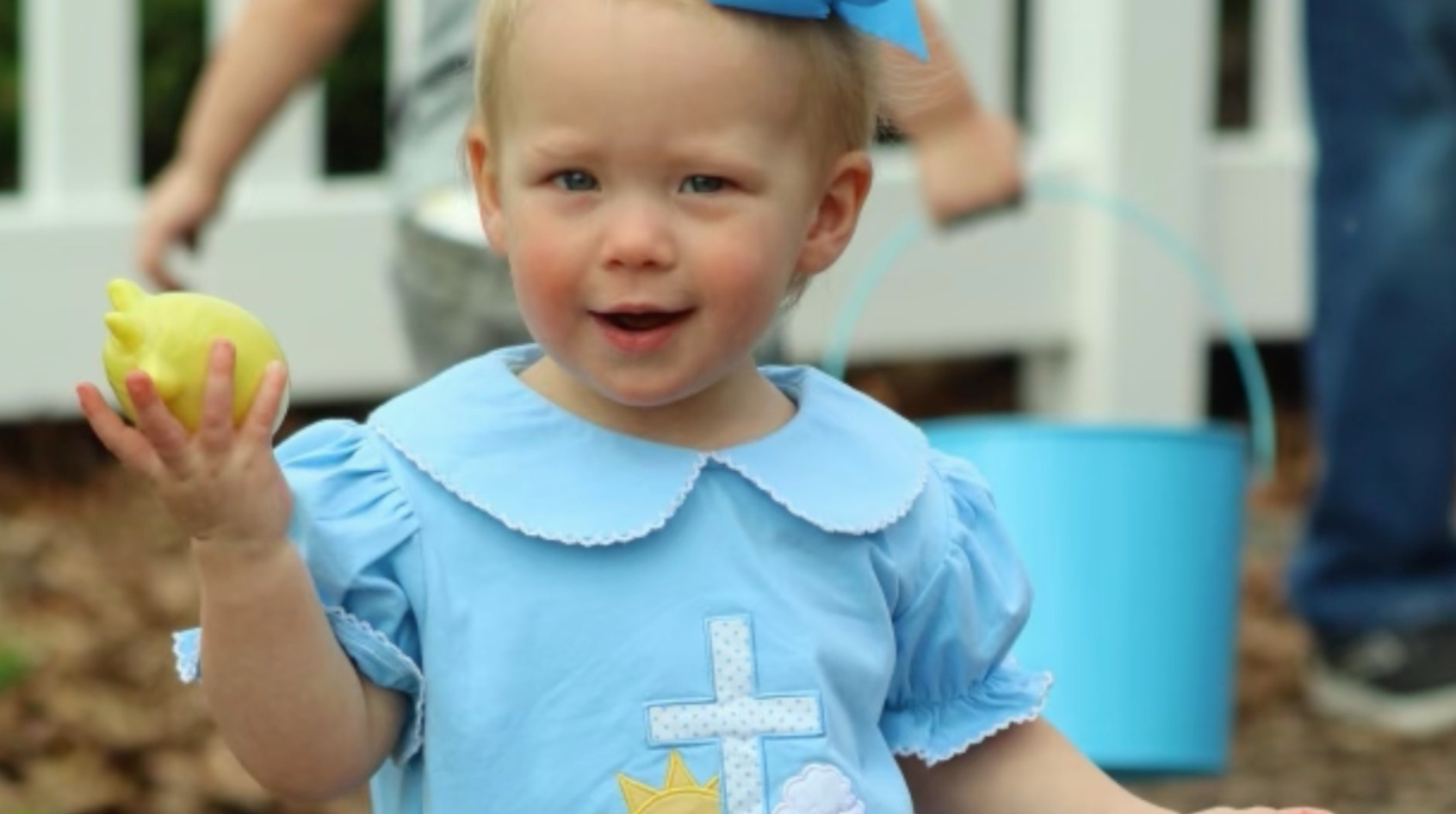 Young child in a light blue dress with a bow, holding a lemon. Outdoors with a blue bucket and fence.