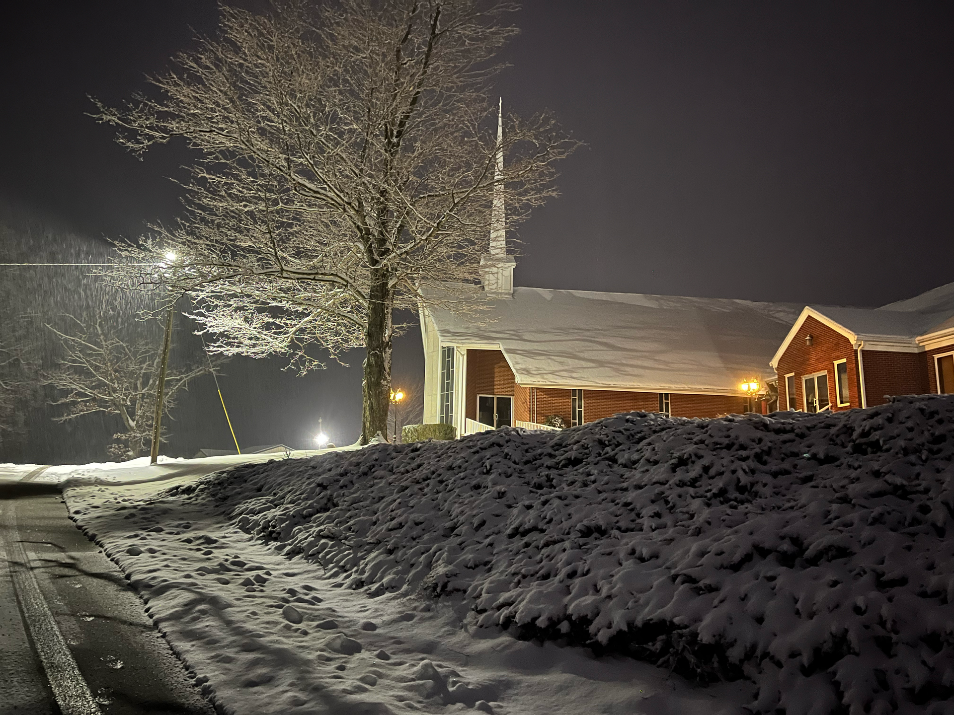 Snow-covered red building with steeple and lit windows at night, next to a snowy road.
