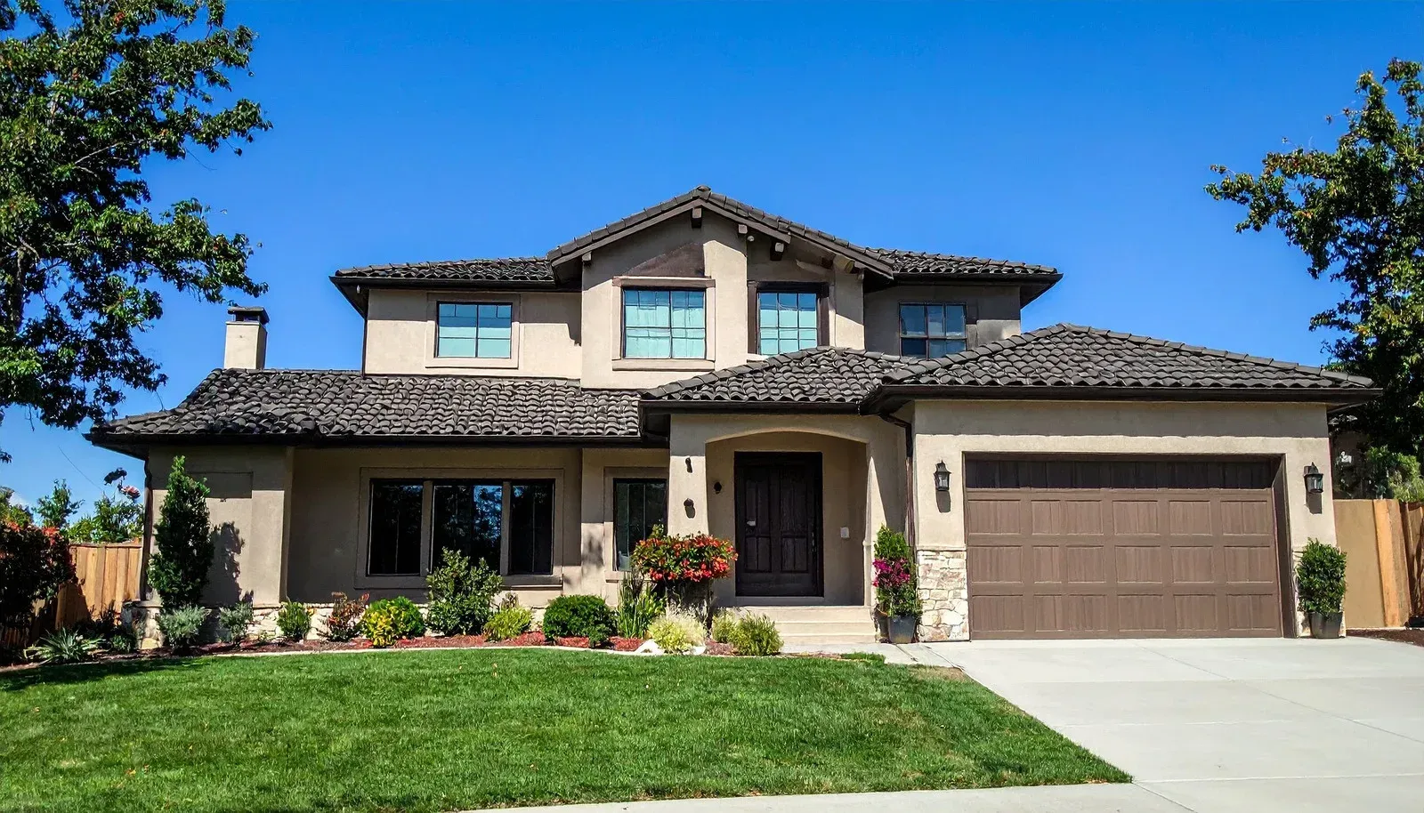 Two-story stucco house with brown roof and garage door; green lawn, blue sky.