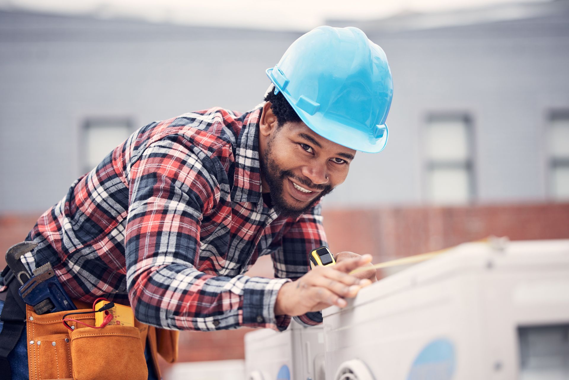 Construction worker in a hard hat and plaid shirt smiling while measuring equipment outdoors.