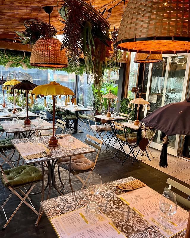 Sala da pranzo del ristorante con tavoli decorati, ombrelloni, piante sospese e lampade intrecciate.