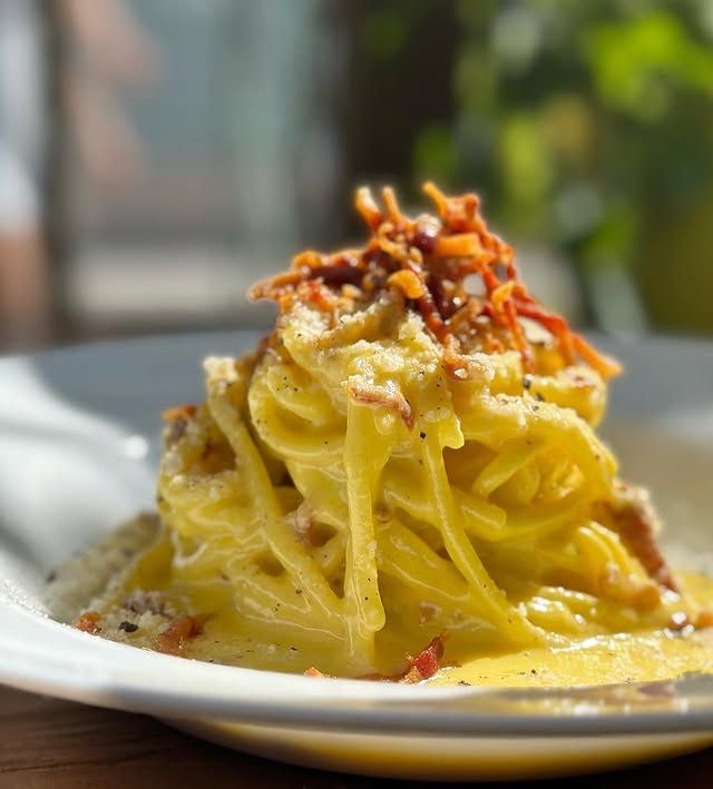 Pasta alla carbonara su un piatto bianco, condita con pancetta croccante e formaggio, salsa gialla brillante.