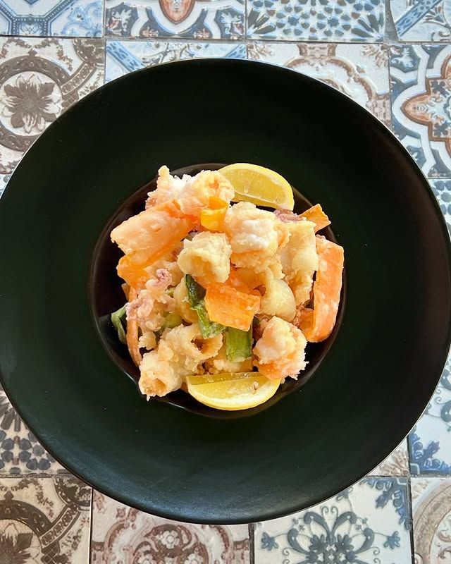 Ciotola di pesce e verdure fritte, guarnita con fette di limone, su un piatto nero.