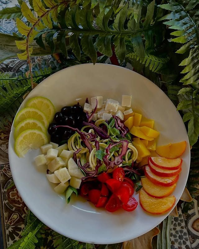 Insalata con tagliatelle di zucchine, fette di frutta e formaggio su un piatto bianco, circondata dal verde.