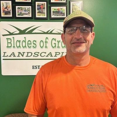 A man wearing glasses and a hat is standing in front of a sign that says blades of grass landscape.