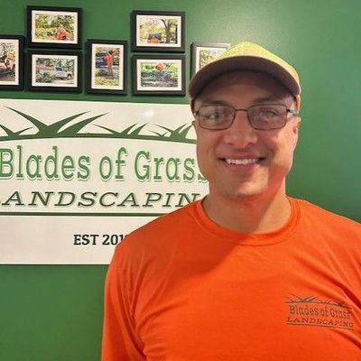 A man wearing glasses and a hat is standing in front of a sign that says blades of grass landscaping.