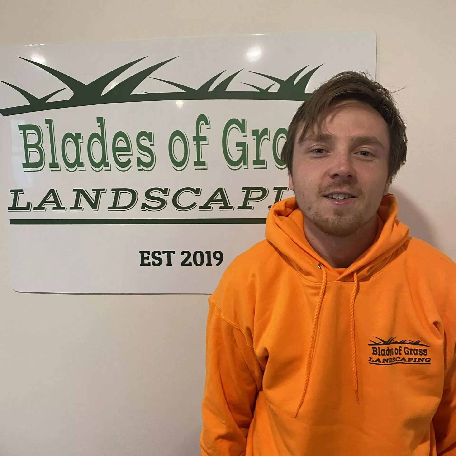 A man in an orange hoodie stands in front of a sign that says blades of grass landscape