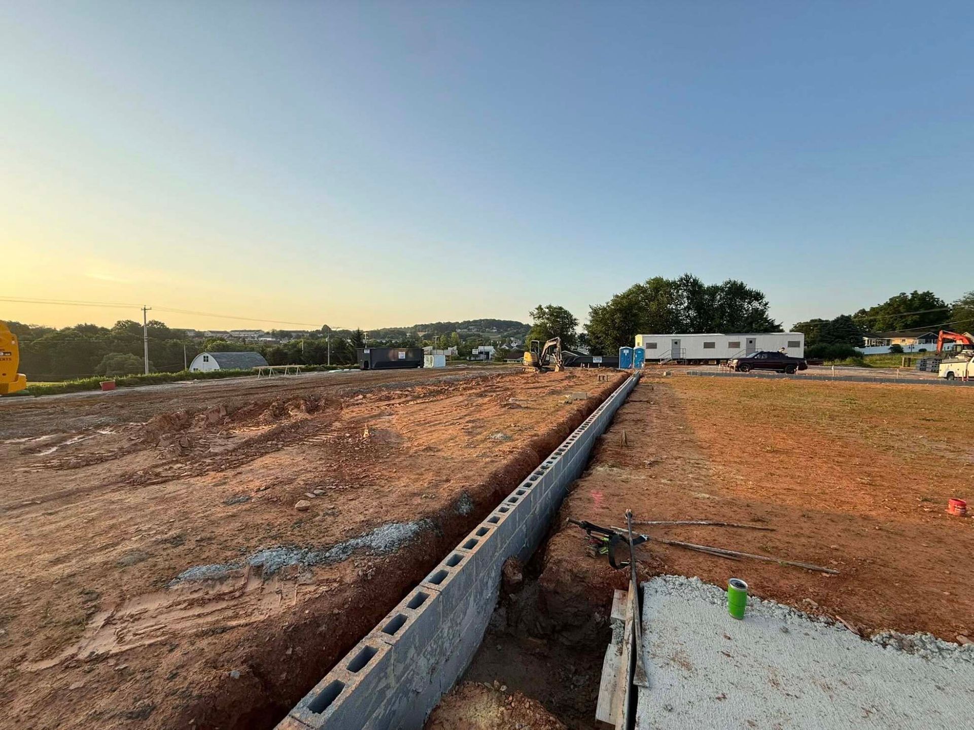 Construction site with a cinder block wall, excavated trenches, and heavy machinery, under a blue sky.