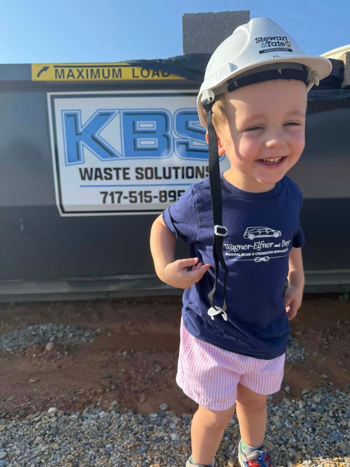 Smiling child in hard hat stands in front of a KBS waste container.