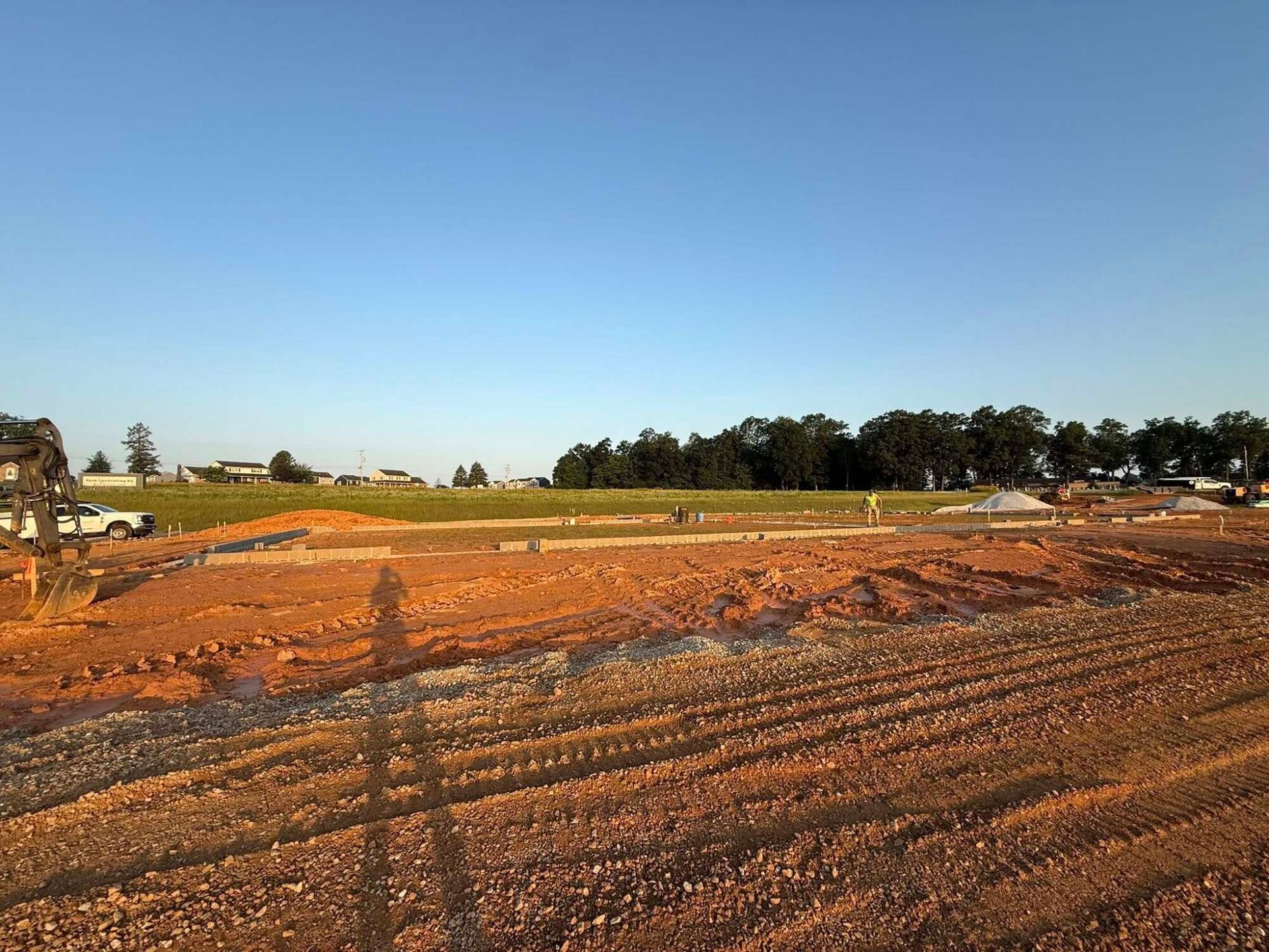 Construction site with red soil, a clear sky, and a line of trees in the background.