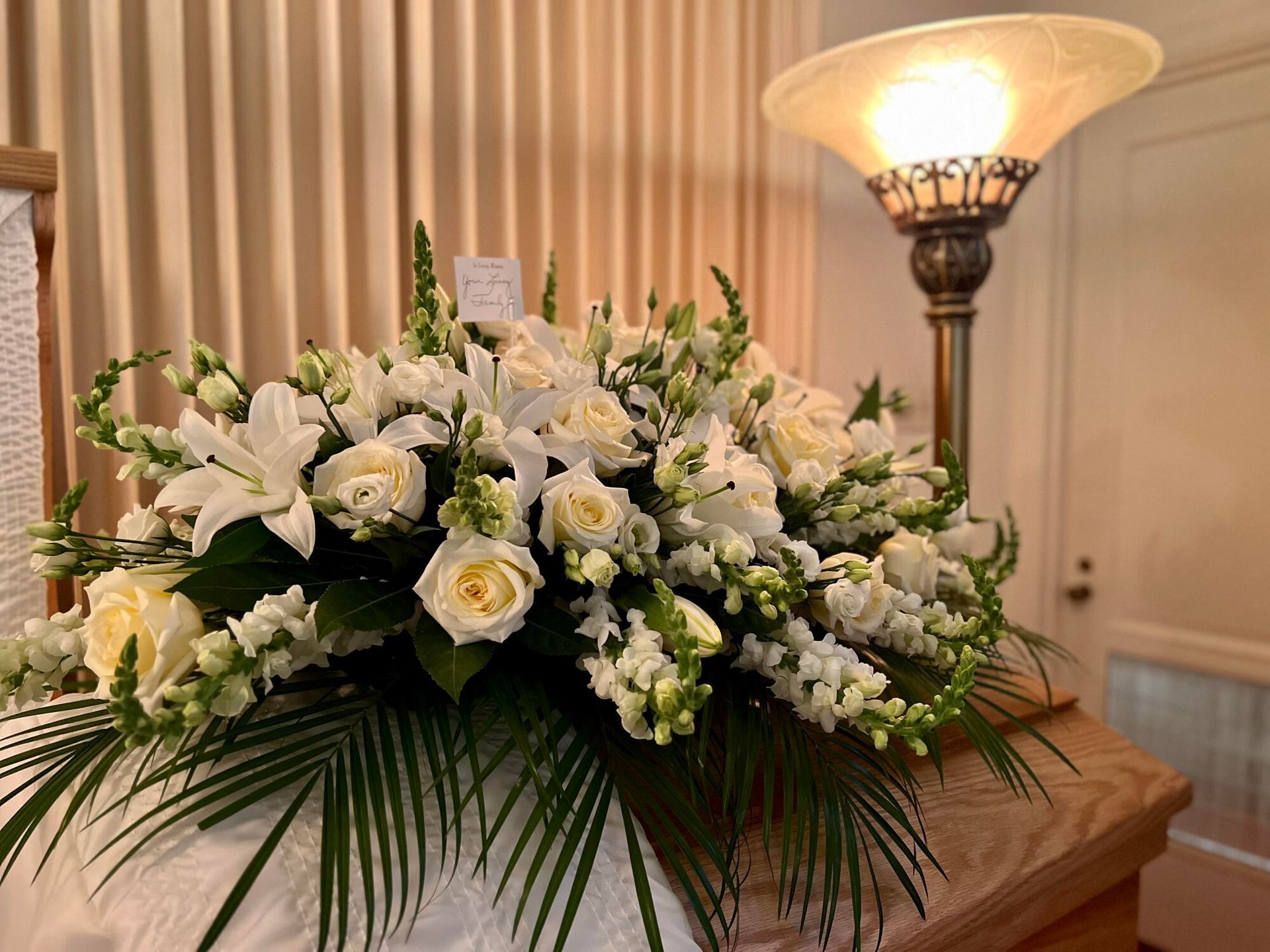 White flower arrangement on a casket, beside a lamp in a room with a light wall.