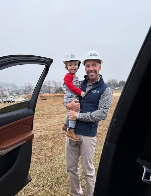 Man holding a child, both wearing white hard hats, standing in a field. Open car door in foreground.