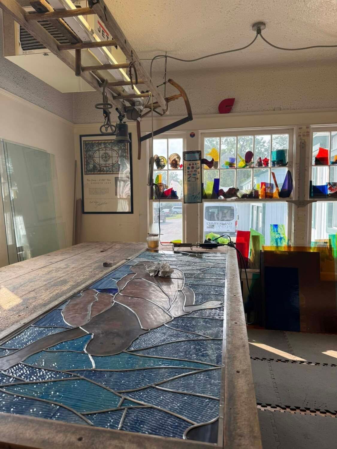 Workshop with a stained glass panel in progress. Windows with colorful glass objects, tools, and a long workbench.