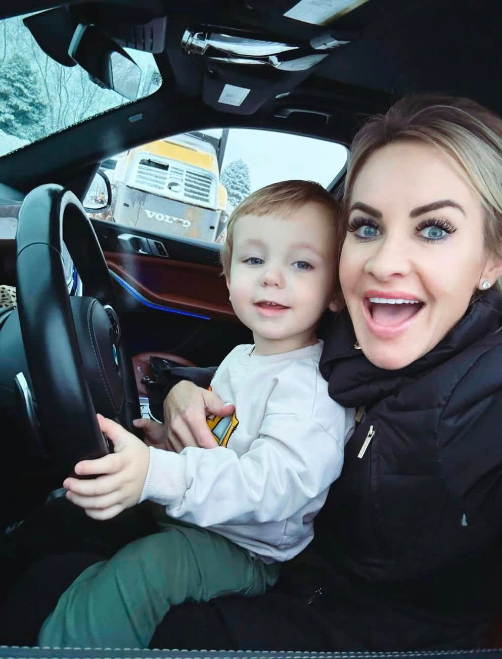 Woman and child smiling inside a car, child holding the steering wheel.